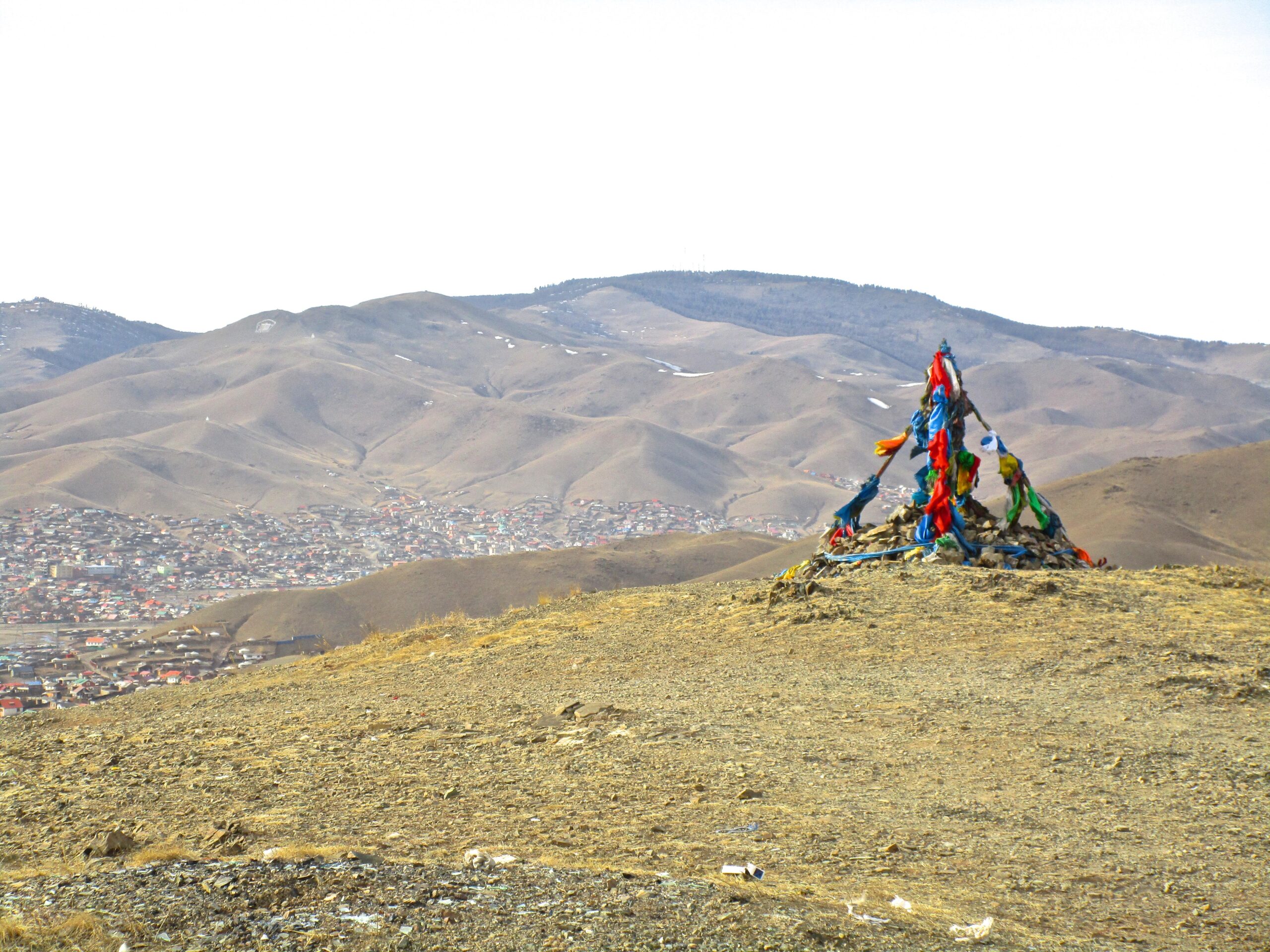 A colorful stone mound, or ovoo, topped with brightly colored fabric ribbons, is situated on a hilltop overlooking a valley with scattered houses and rolling mountains in the background. The sky is light and clear, emphasizing the natural landscape. District 15 Network mountain bike trail.