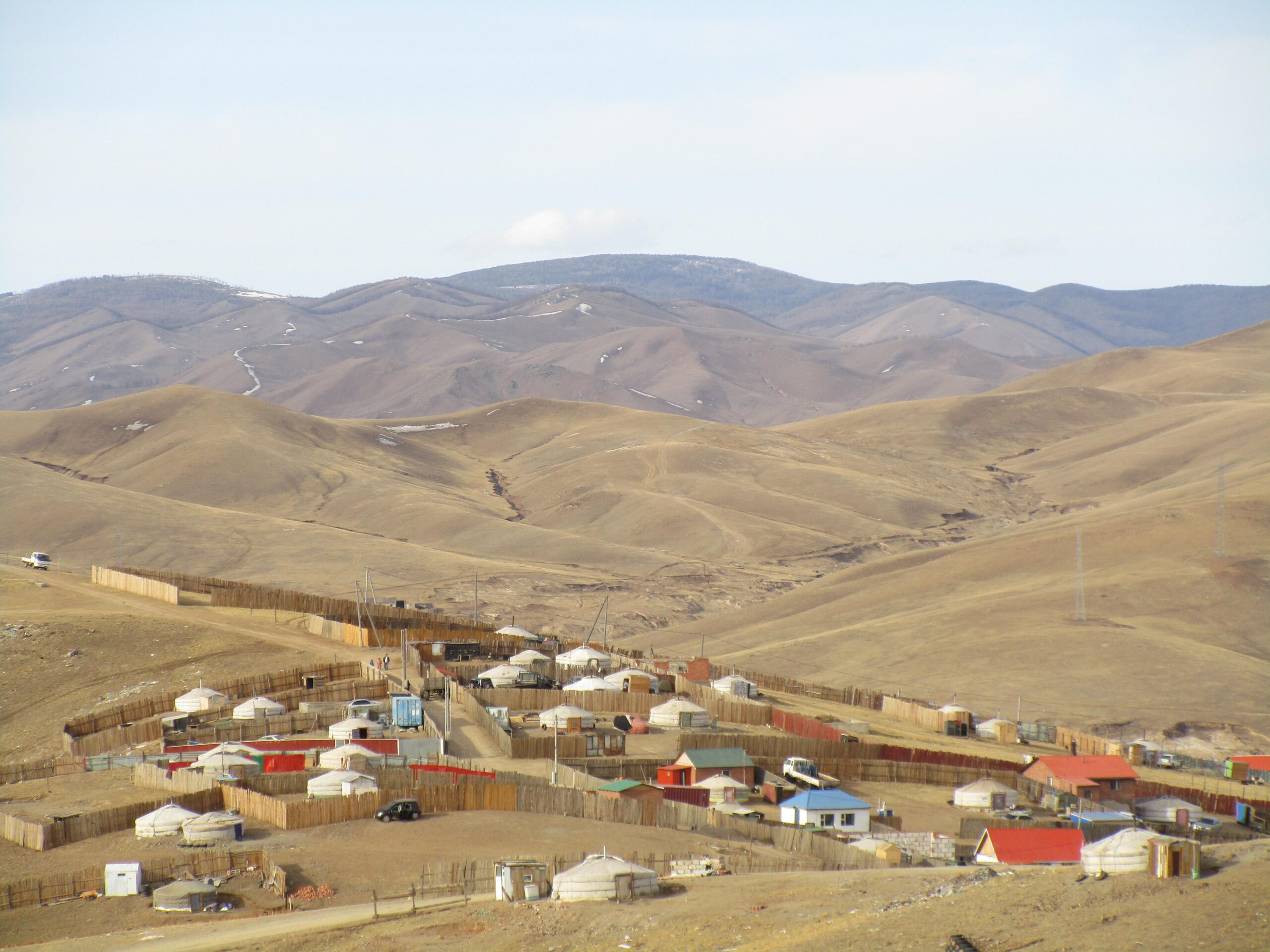 Landscape featuring rolling hills in the background and a settlement with traditional yurts and wooden structures in the foreground. The area is arid with sparse vegetation, and a few vehicles are visible along the dirt paths. The sky is partly cloudy. District 15 Network mountain bike trail.