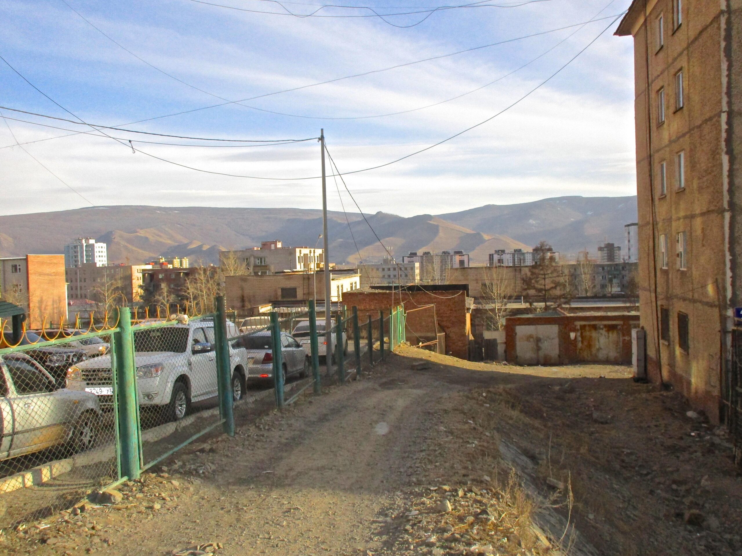 Dirt road winding between buildings, with parked cars along a fenced area; mountains visible in the background under a blue sky. District 15 Network mountain bike trail.