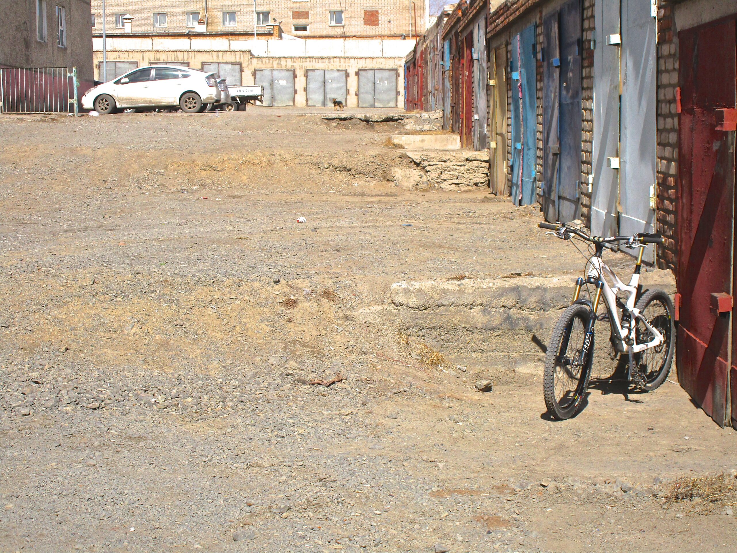 A gravel alleyway with a white bicycle resting against a concrete step. In the background, there are various colored garage doors and a parked white car. The ground is dry and dusty, with patches of dirt and small rocks. District 15 Network mountain bike trail.