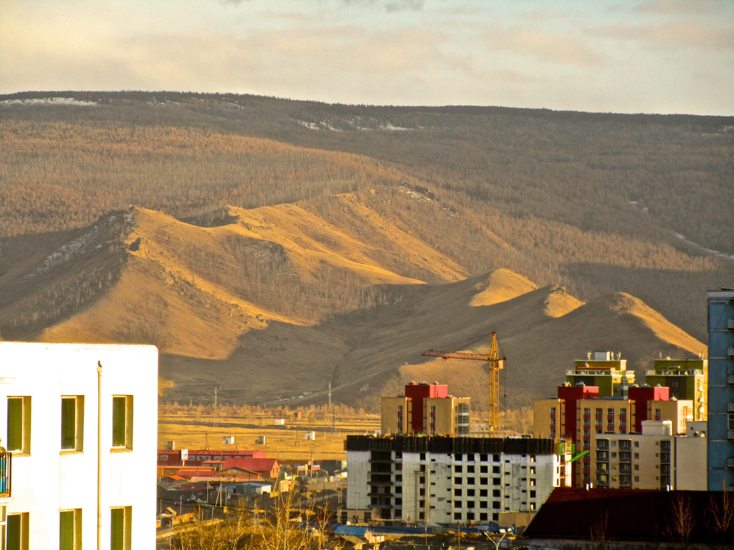 A view of rolling hills and mountains in the background, with a mix of natural landscapes and urban buildings in the foreground. The scene features a construction crane and various structures, including residential and commercial buildings, under a sky with soft clouds and warm sunlight illuminating the landscape. The Beast mountain bike trail.