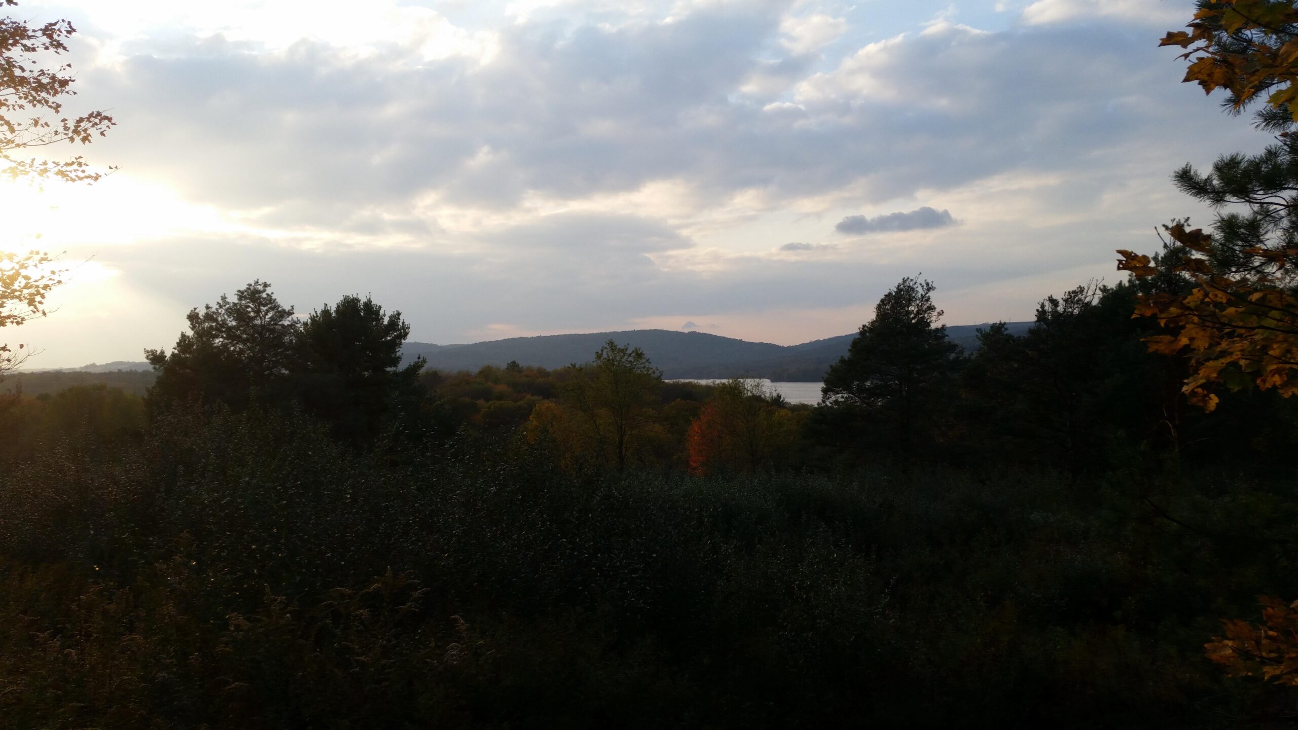 A scenic landscape featuring a hillside covered with trees and shrubs, under a cloudy sky. The sun is setting, casting warm light over the scene, and a river can be seen in the background, bordered by more hills. Autumn foliage adds hints of orange and yellow to the greenery, creating a tranquil and peaceful atmosphere. Yellow Creek State Park mountain bike trail.