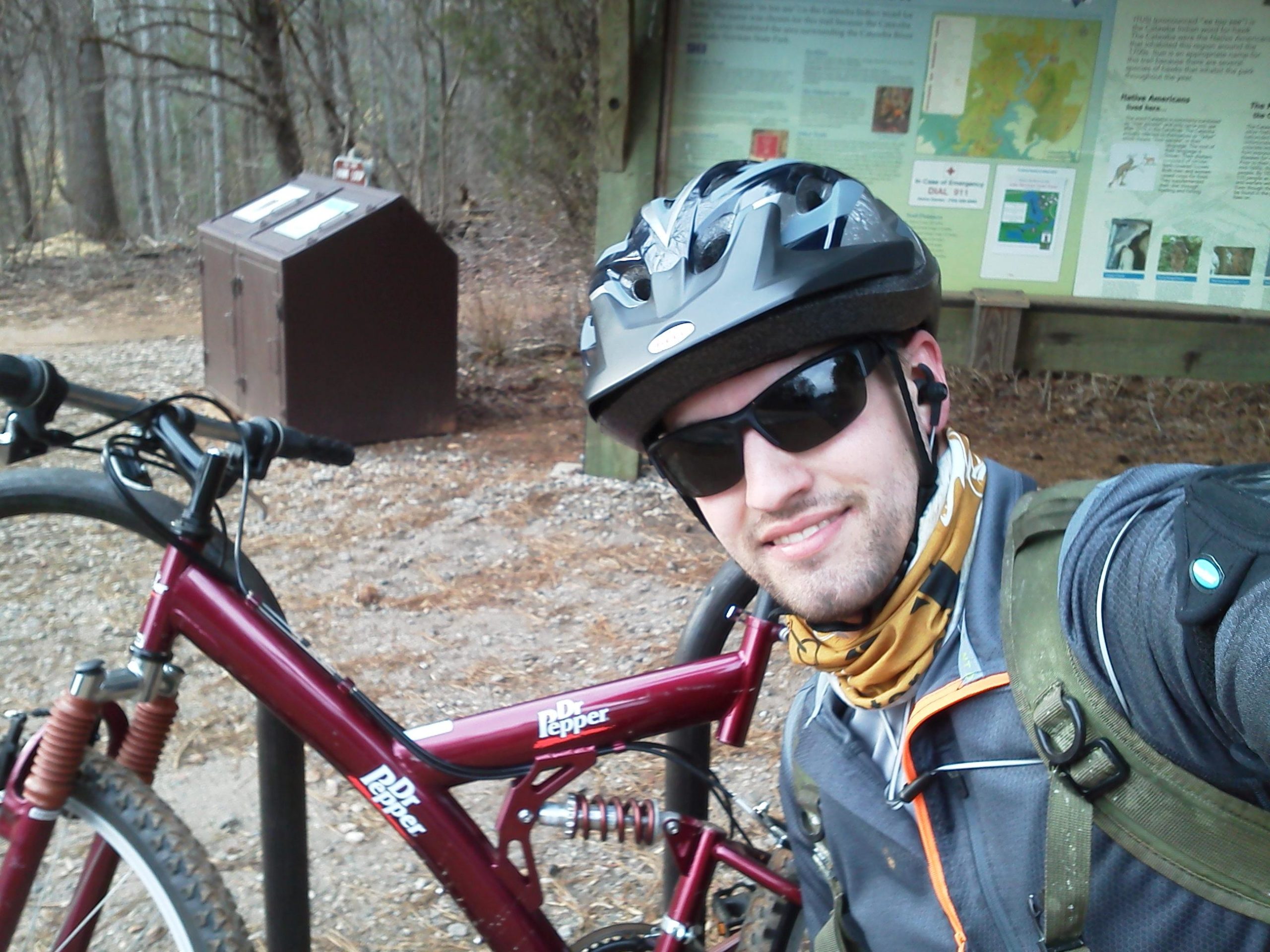 A person wearing a helmet and sunglasses poses for a selfie next to a maroon mountain bike. They are dressed in a jacket and a neck gaiter, with a backpack visible on their shoulder. In the background, there is a information board with a map and text, and a brown storage box. The setting is in a wooded area with gravel ground. Itusi @ Lake Norman State Park mountain bike trail.