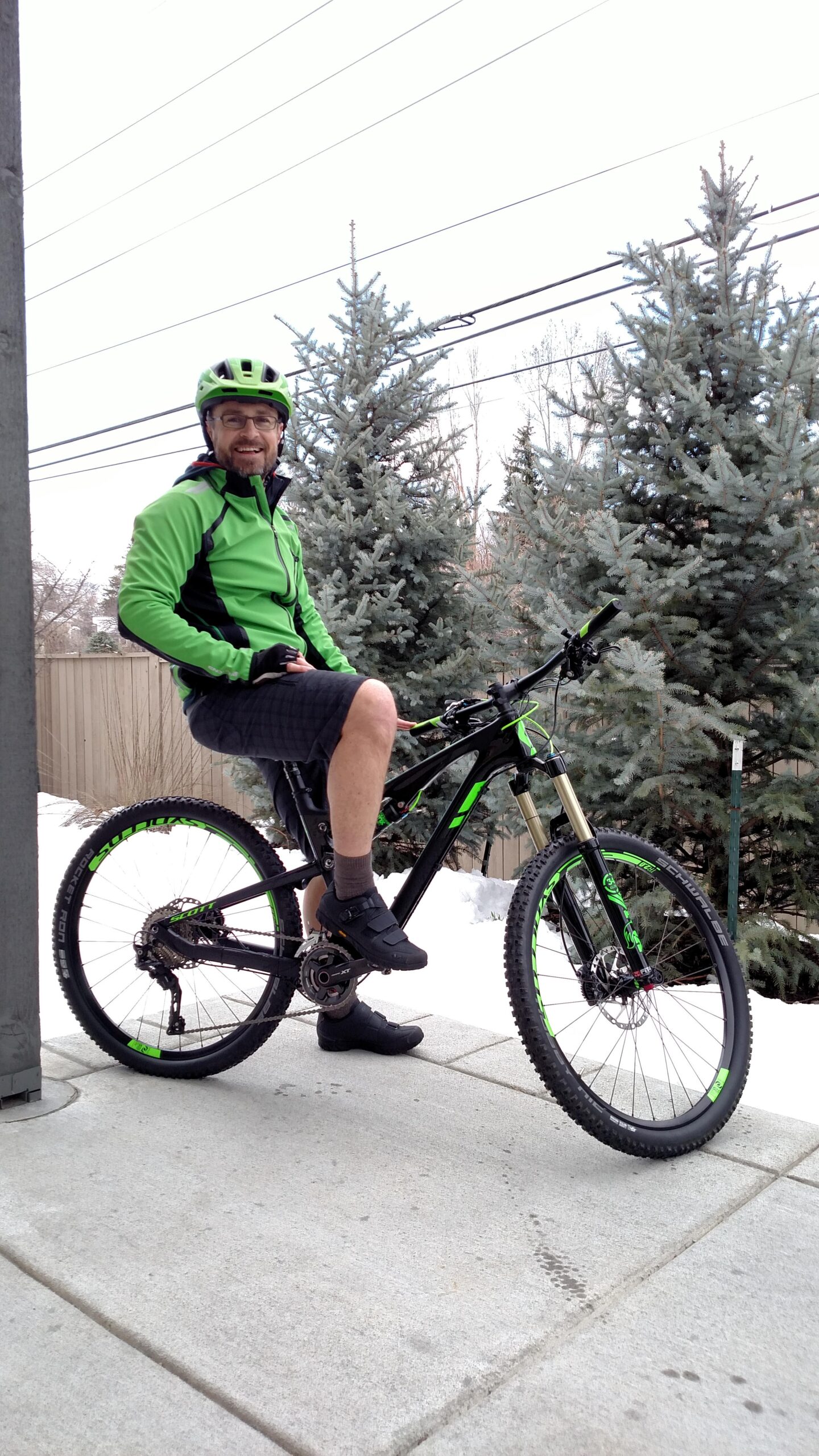 Scott Genius 710: A man wearing a bright green cycling jacket and helmet sits on a mountain bike, smiling at the camera. He has a short beard and is wearing dark shorts and cycling shoes. The background features snow on the ground and evergreen trees. Power lines are visible in the sky above.