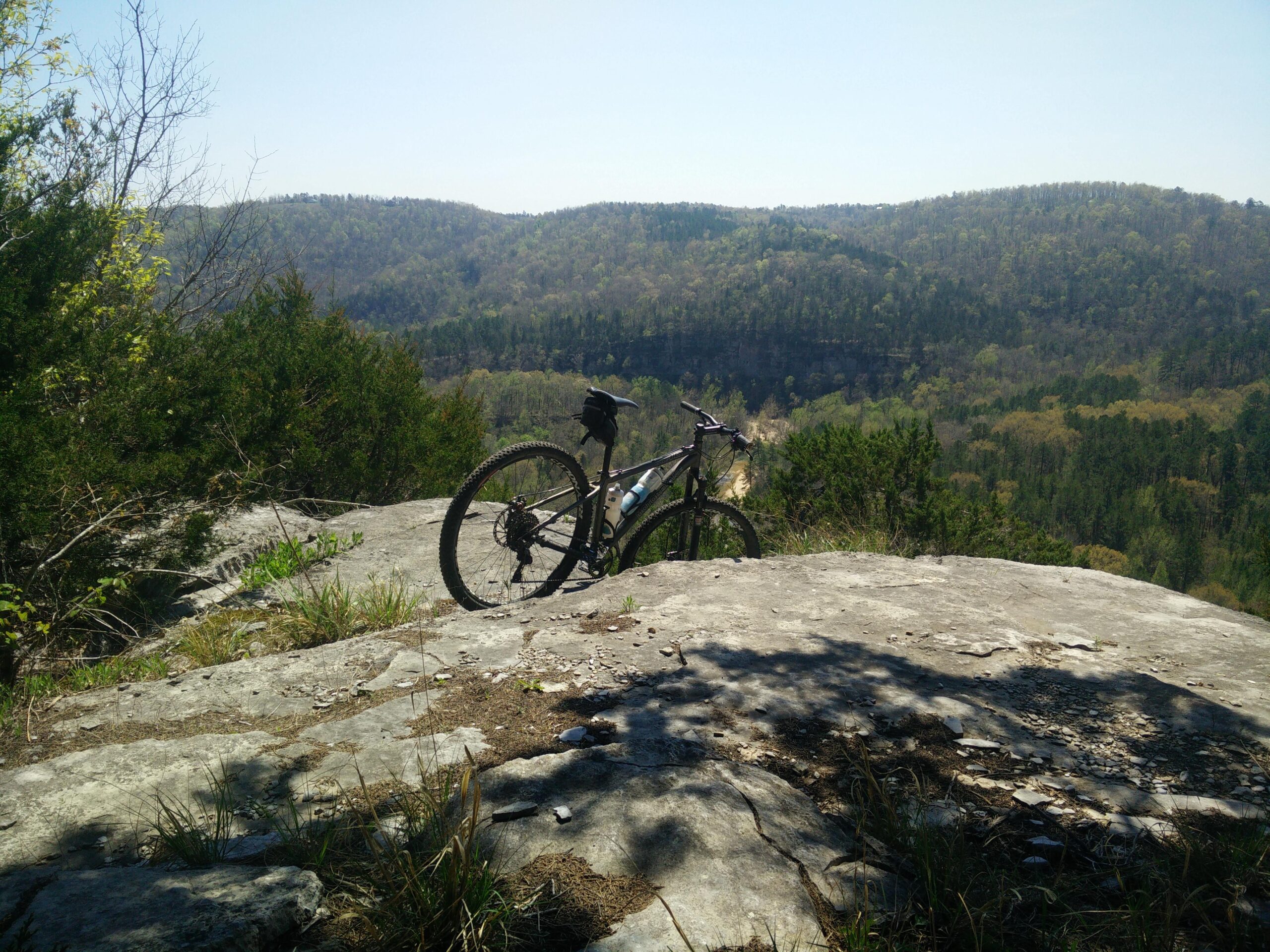 A mountain bike parked on a rocky outcrop overlooking a scenic view of rolling hills and trees under a clear blue sky. The foreground features rocky terrain with patches of grass and small plants, while the background displays lush greenery in various shades, indicating a vibrant landscape. Syllamo Trails mountain bike trail.