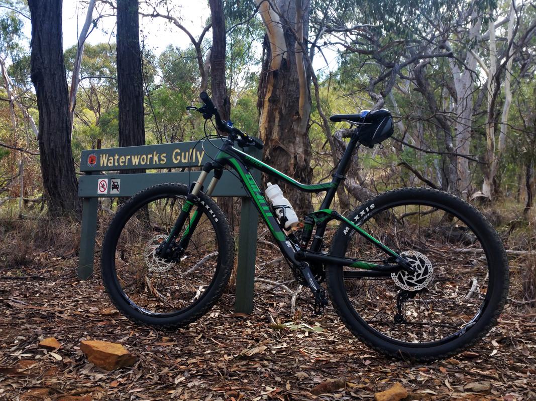 Giant Stance 27.5 2: Mountain bike leaning against a "Waterworks Gully" sign surrounded by trees and foliage, with rocky ground and dry leaves visible.