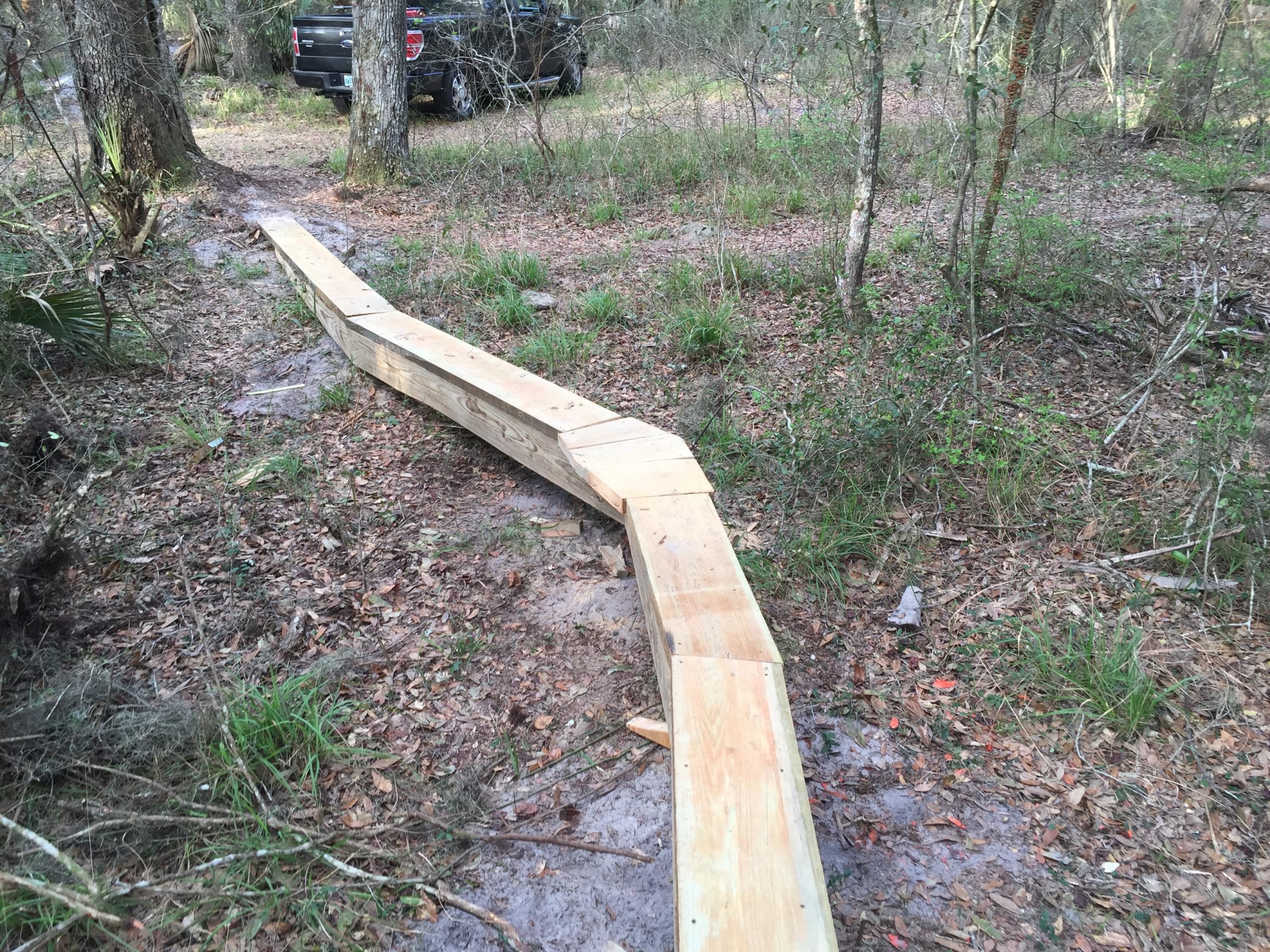 A wooden walkway curving through a wooded area, surrounded by grass and fallen leaves, with a black vehicle parked in the background. The path is designed to navigate the uneven terrain in a natural setting. Nocatee mountain bike trail.