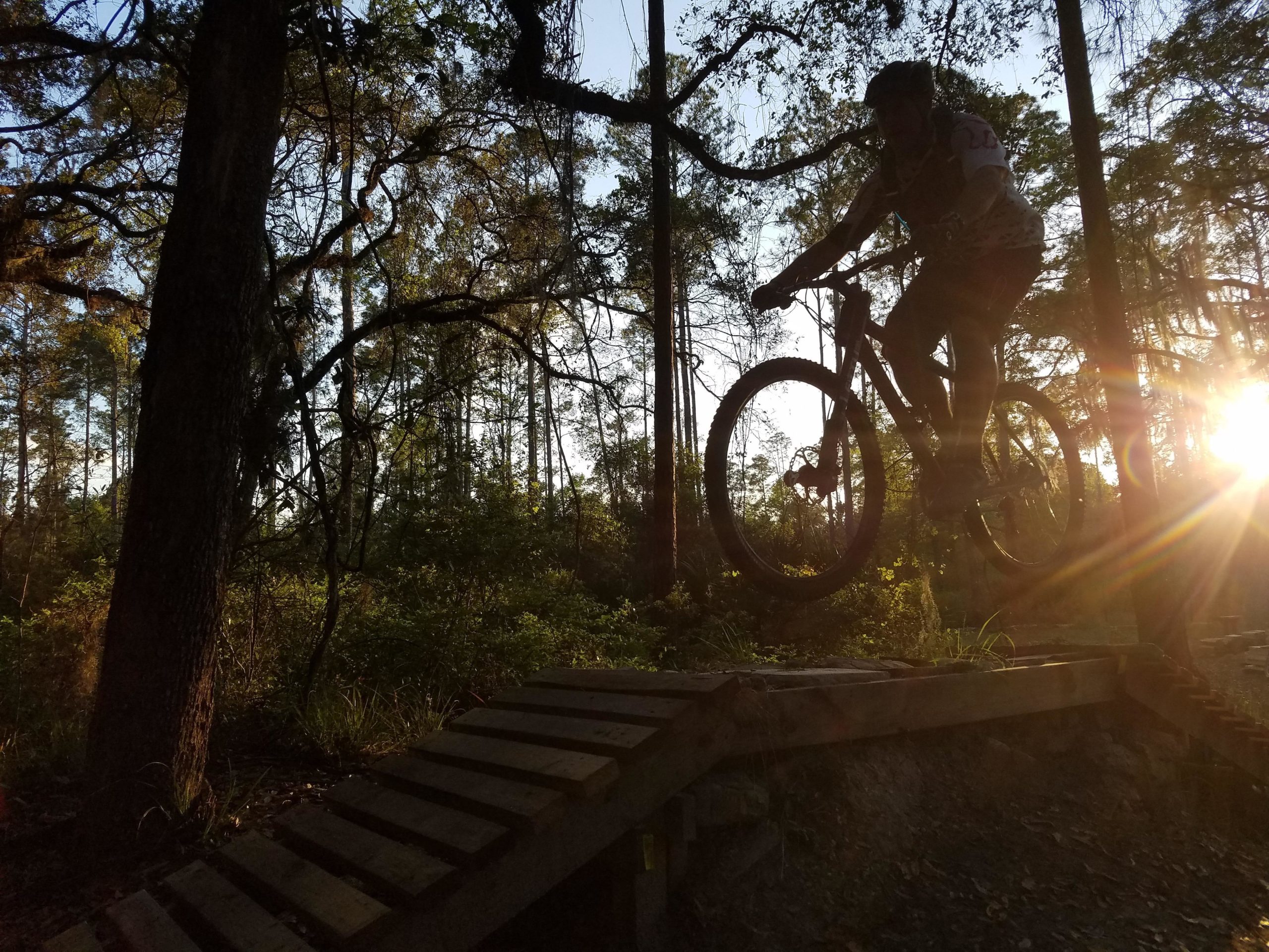 A mountain biker is captured mid-air while jumping off a wooden ramp in a forested area at sunset. The silhouette of the rider is framed against the warm glow of the setting sun, with trees and greenery surrounding the scene. Nocatee mountain bike trail.