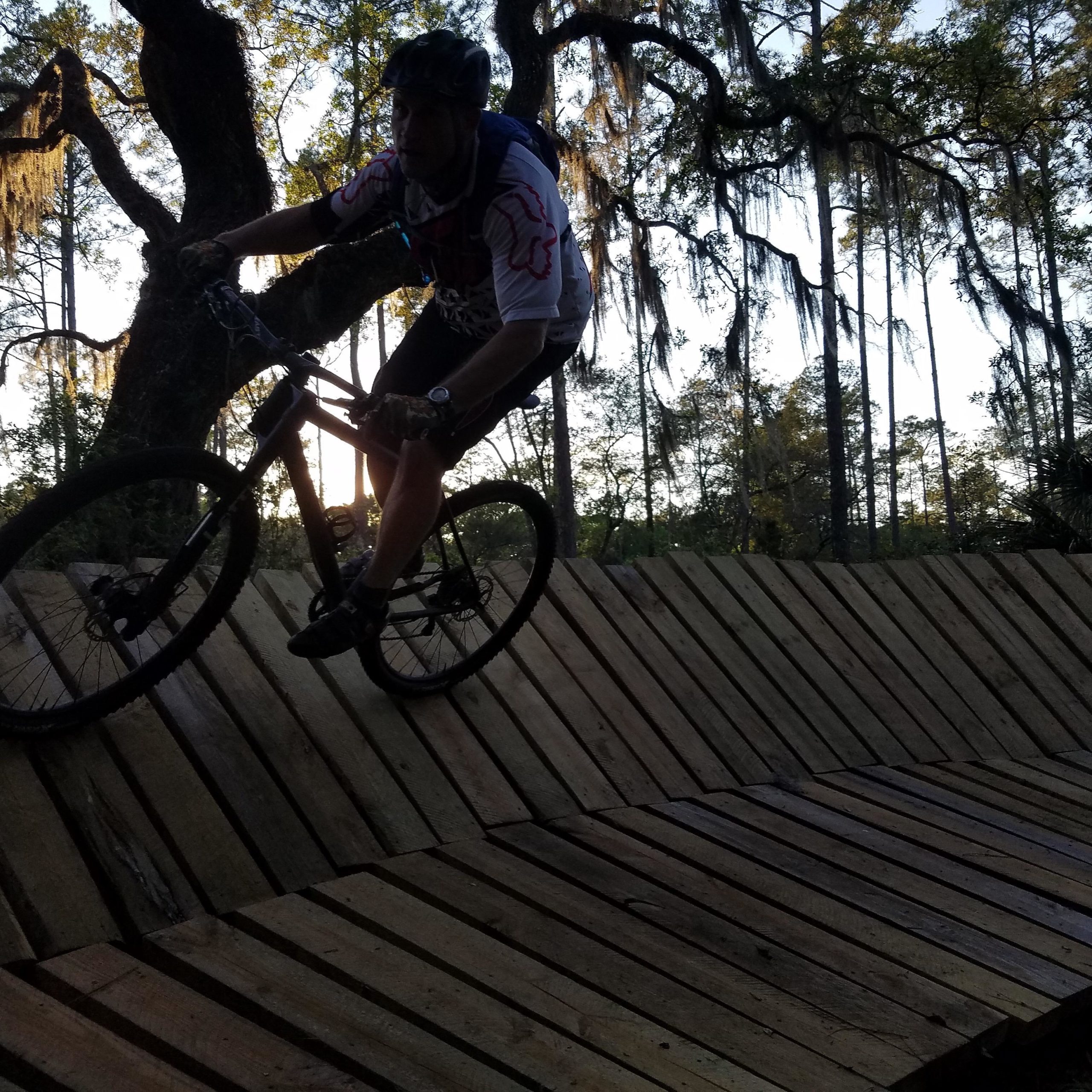 A mountain biker navigating a wooden ramp in a forested area during sunset, with trees and Spanish moss in the background. Nocatee mountain bike trail.