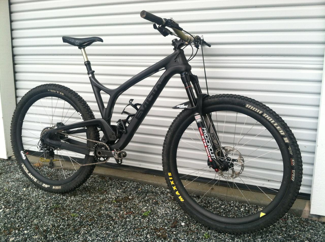 Evil The Following: A black mountain bike leaning against a textured wall, featuring a sleek frame, wide tires, and suspension forks. The bike is positioned on a gravel surface, showcasing its professional-grade components, including a drivetrain and disc brakes.