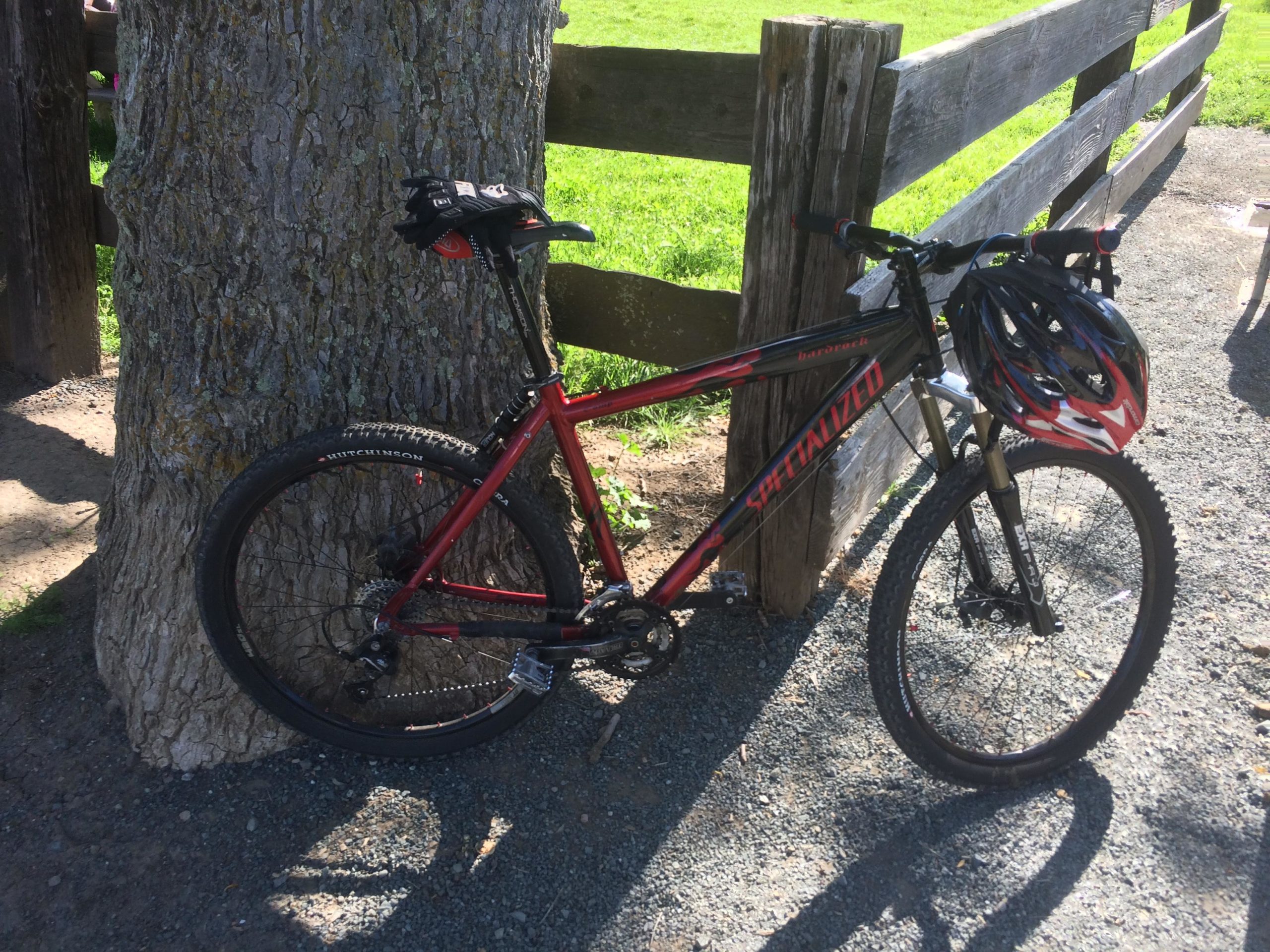 A red and black mountain bike leaning against a large tree, with a helmet resting on the handlebars. The background features a wooden fence and a grassy area, illuminated by sunlight. Black Diamond Mines mountain bike trail.