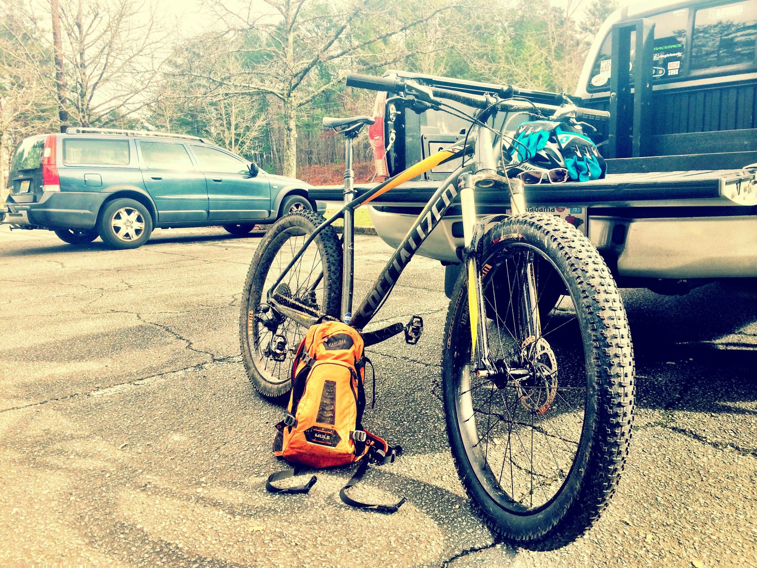Specialized Fuse Comp 6Fattie: A mountain bike with a black and orange frame is leaning on the ground next to an orange backpack. In the background, two vehicles are parked on a cracked asphalt surface, surrounded by trees. The scene captures a moment before an outdoor biking adventure.