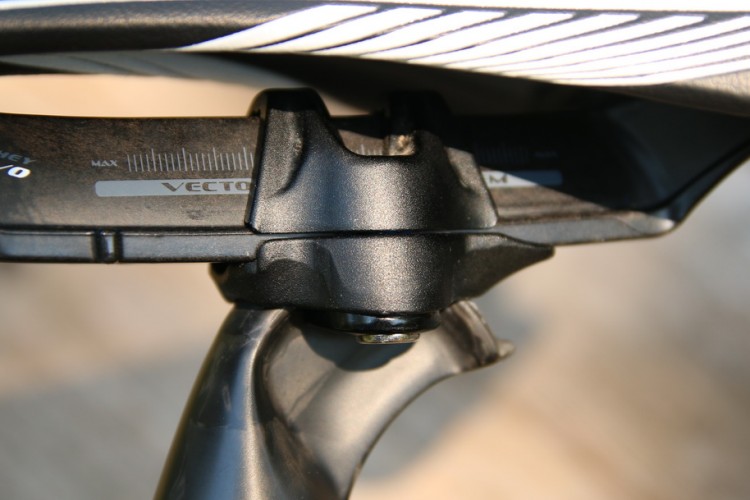 Close-up of a bicycle saddle's adjustment mechanism, featuring a scale for measuring height with labels including "MAX" and "VECTOR EVO." The image highlights a black mounting bracket connected to the seat post.