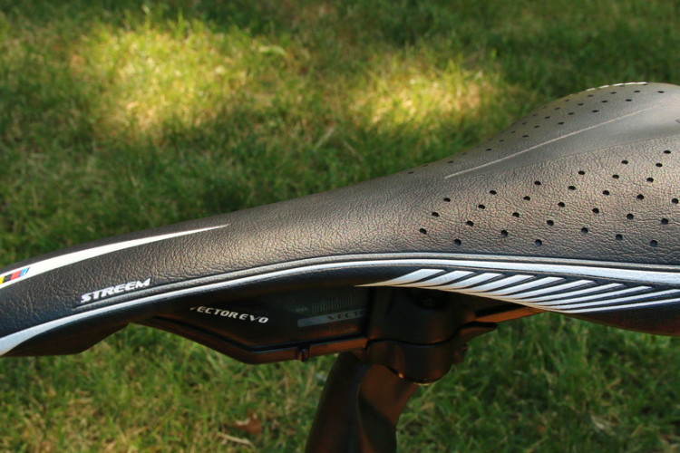 Bicycle saddle with a sleek black design, featuring perforations for ventilation, mounted on a seat post against a grassy background.