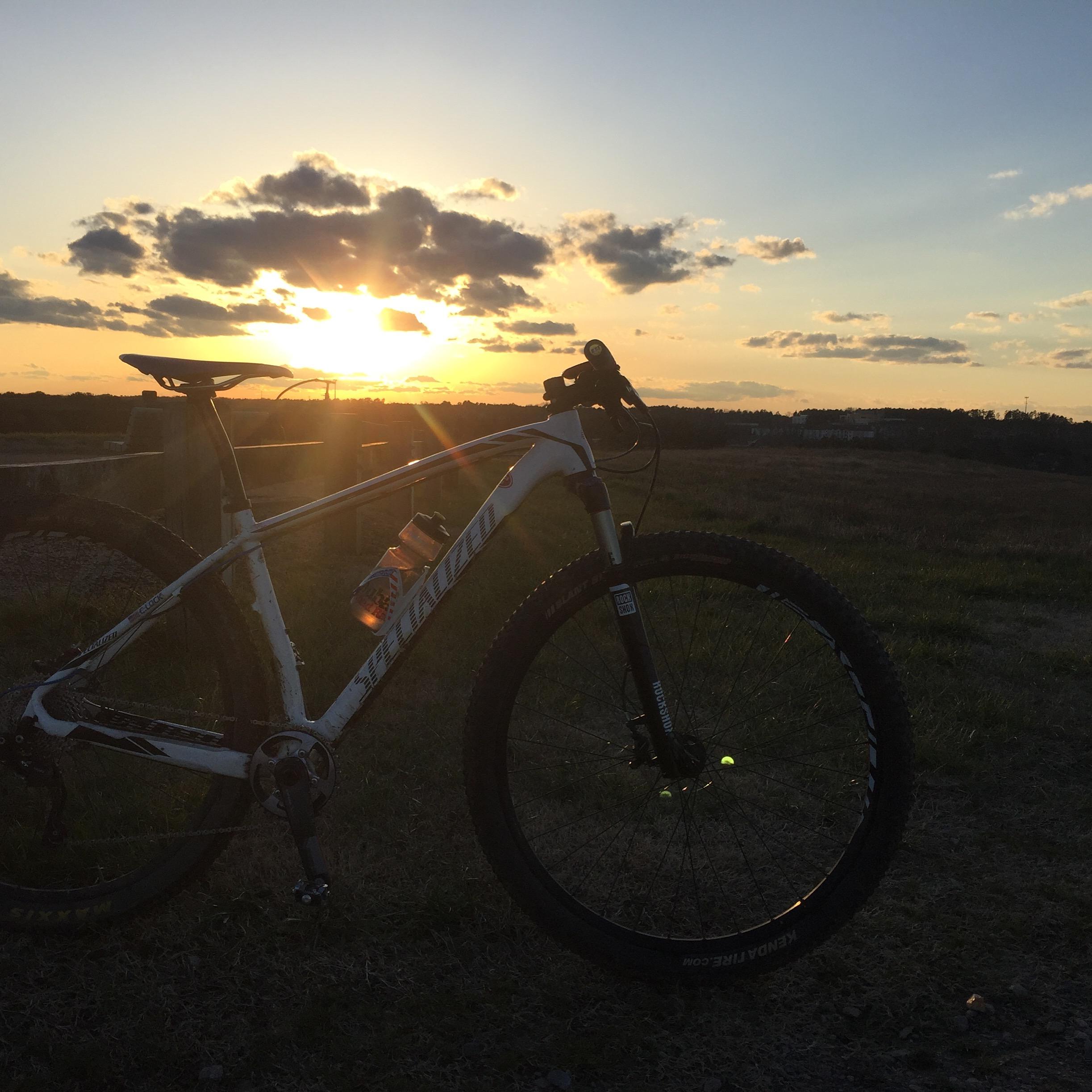 A mountain bike is positioned in the foreground, silhouetted against a sunset sky filled with vibrant colors and clouds. A water bottle is attached to the bike, and the sun