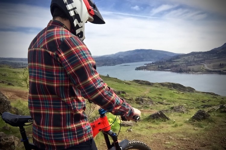 A mountain biker wearing a plaid shirt and helmet stands next to their bike, overlooking a scenic landscape with a lake and rolling hills in the background. The sky is partly cloudy, and the terrain is green and rocky, suggesting an outdoor adventure.