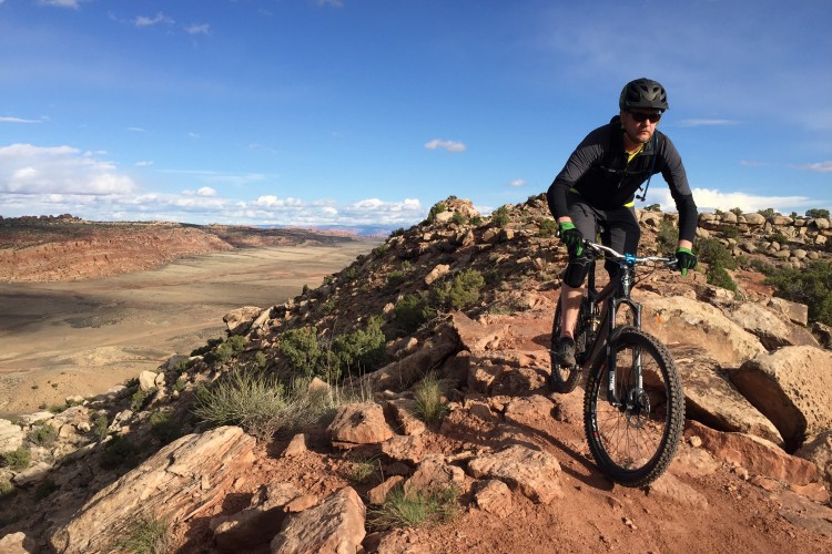 A mountain biker navigating a rocky trail with a panoramic view of a desert landscape under a partly cloudy sky. The cyclist is wearing a helmet and cycling attire, positioned on a sloped terrain surrounded by boulders and sparse vegetation.
