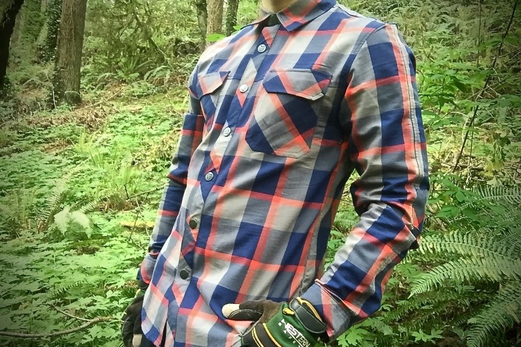 A person wearing a plaid button-up shirt stands confidently in a lush green forest setting, surrounded by ferns and trees. Their gloved hand is resting on their hip, and the shirt features shades of blue, gray, and red in a checkered pattern.