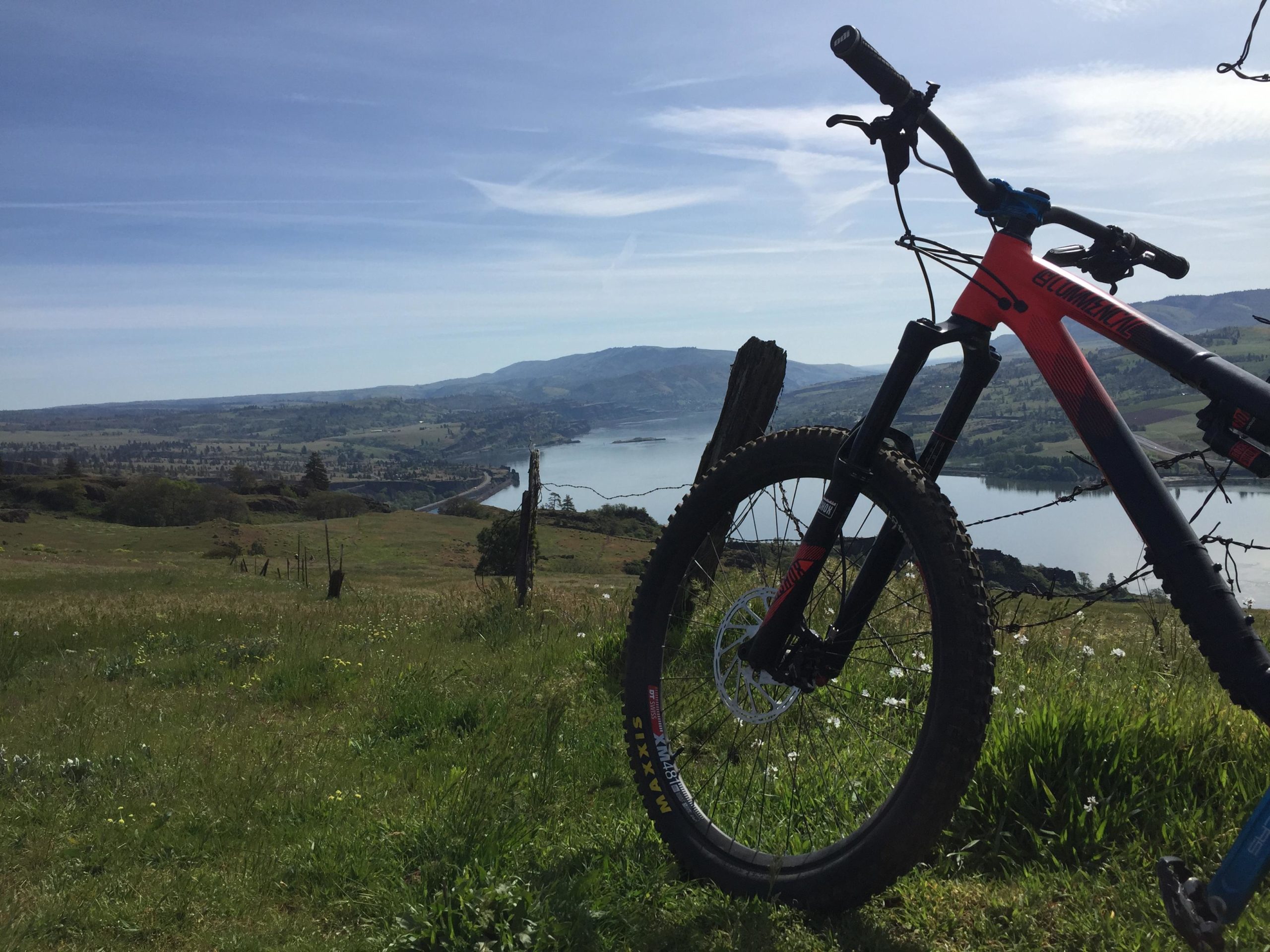 A mountain bike leaning against a fence in a lush green landscape overlooking a river and distant hills under a blue sky with wispy clouds. Syncline mountain bike trail.