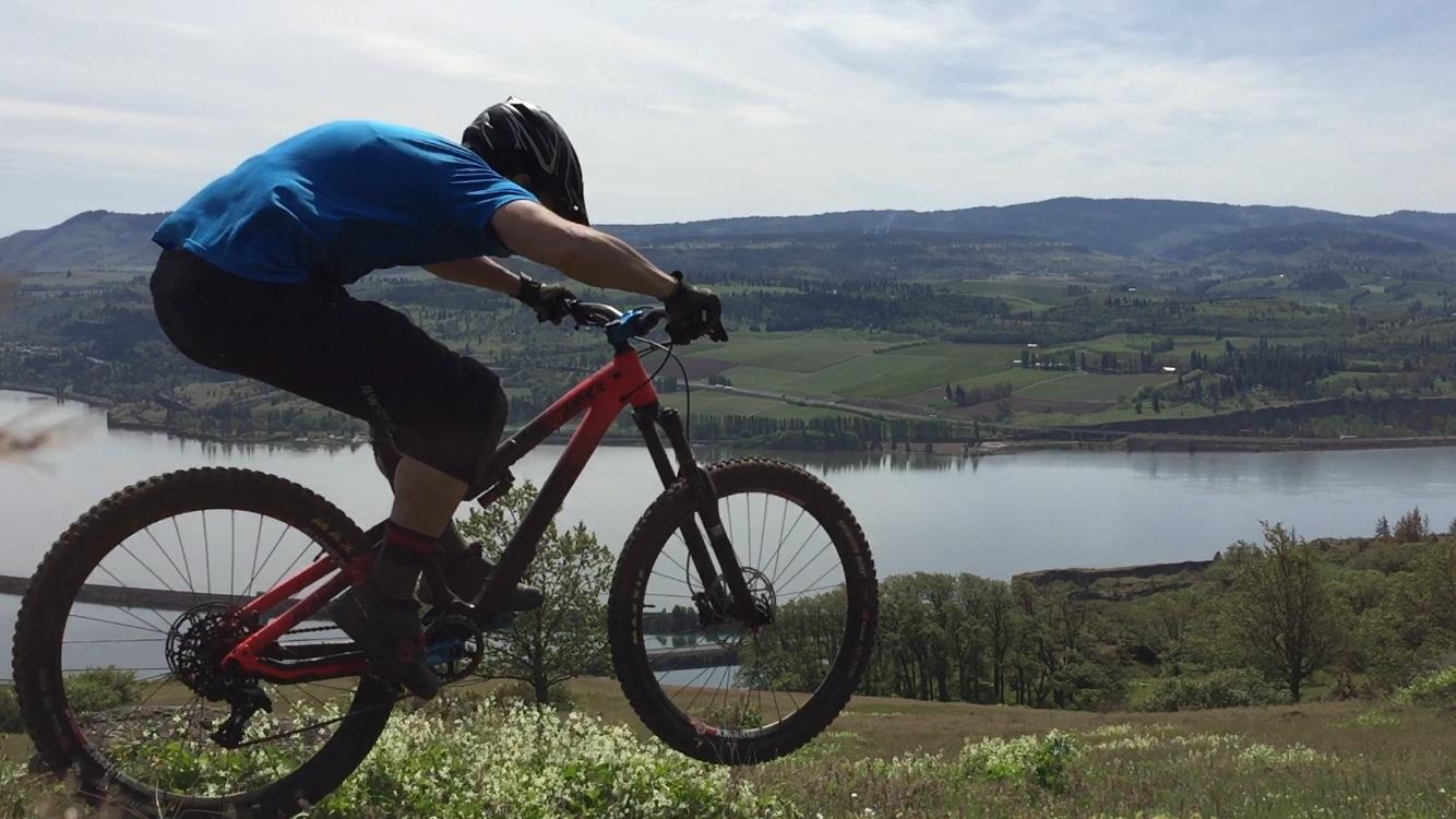 A mountain biker in a blue shirt and helmet is performing a jump over a grassy hill, with a scenic background of rolling hills and a river. The sky is clear, and the landscape features lush greenery and agricultural fields. Syncline mountain bike trail.