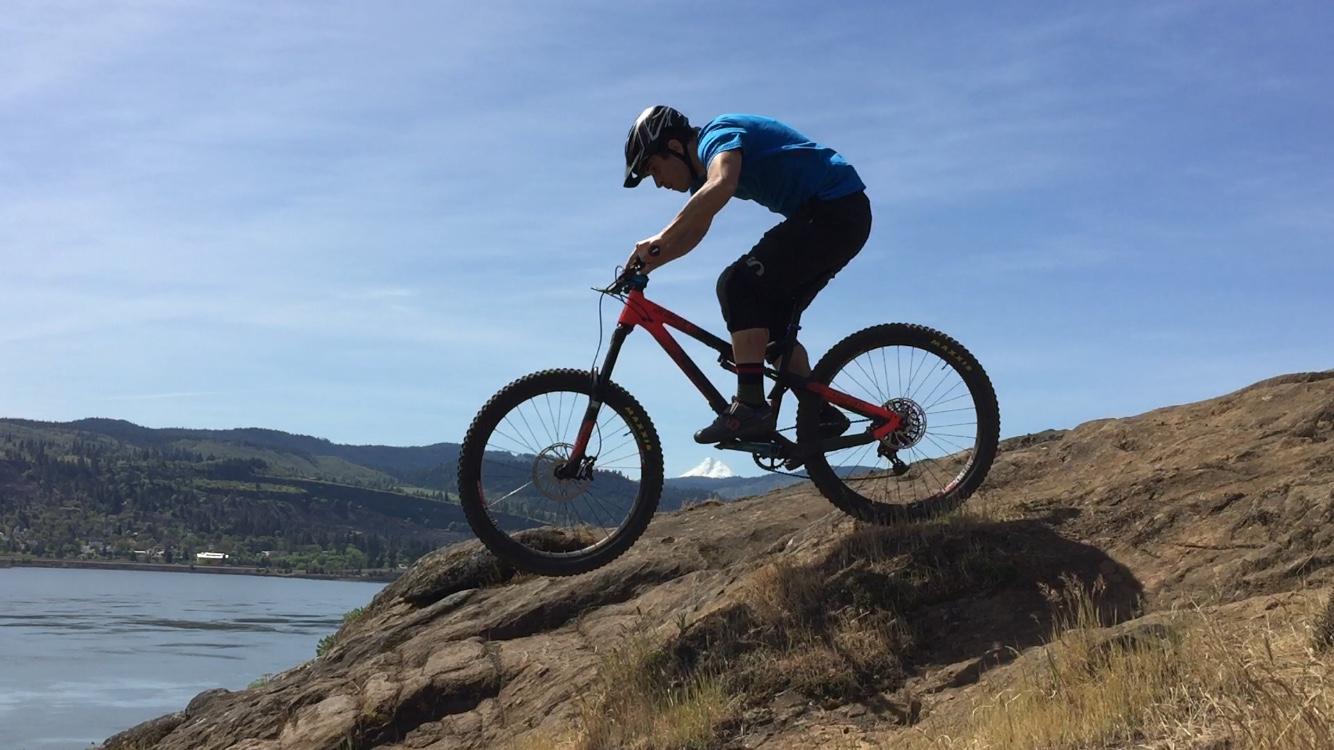A mountain biker in a blue shirt and helmet descends a rocky cliff, with one wheel off the ground, against a scenic backdrop of hills and a river under a clear blue sky. Syncline mountain bike trail.