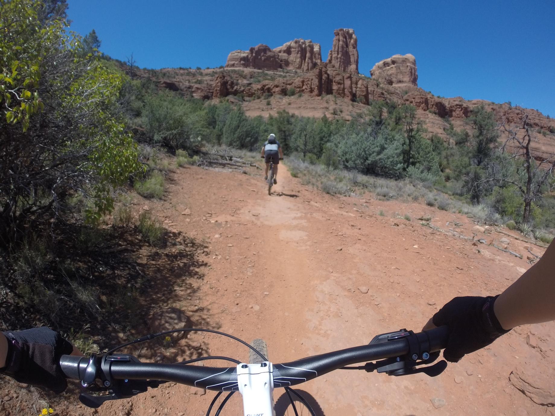 A mountain biker rides on a dusty trail surrounded by greenery, with sandstone formations in the background under a clear blue sky. The view is from the cyclist's perspective, showing the handlebars and part of the bike. Mescal Trail mountain bike trail.
