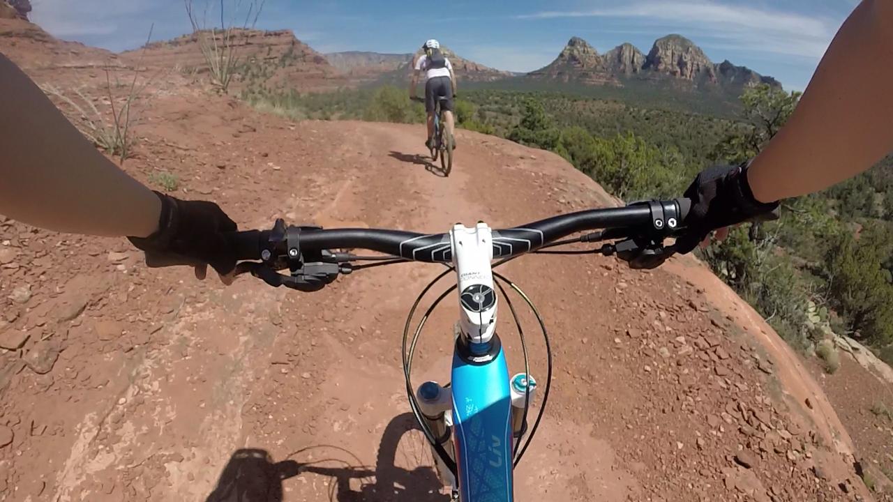 Mountain biking scene from a first-person perspective, showing the handlebars of a bike on a rocky trail with a rider in the background and scenic red rock formations in the distance. Mescal Trail mountain bike trail.