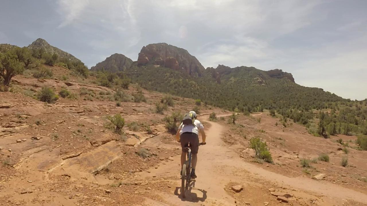 A person riding a mountain bike on a dirt trail surrounded by rocky terrain and sparse vegetation, with mountains in the background under a sunny sky. Chuck Wagon mountain bike trail.