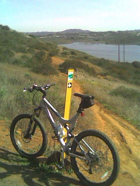 Specialized Stumpjumper: A mountain bike rests on a dirt path surrounded by green hills, with a lake visible in the background. A trail marker stands nearby, indicating the direction for cyclists. The scene is bright and sunny, showcasing a peaceful outdoor setting.