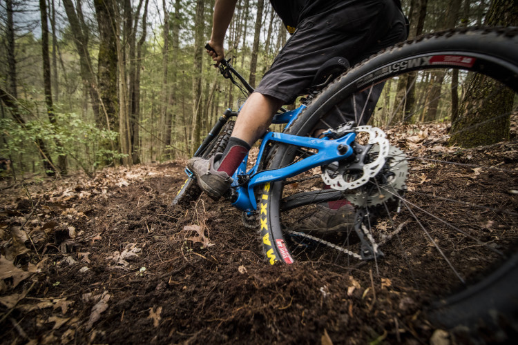 A mountain biker navigating a muddy trail in a forest, with focus on the bike's blue frame, tires, and the rider's legs and footwear as they maneuver through the terrain.