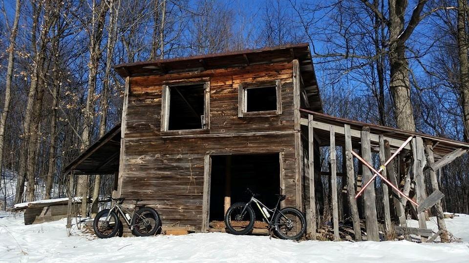 Alt text: A rustic wooden cabin with a slanted roof, partially covered in snow, surrounded by bare trees under a clear blue sky. Two bicycles are leaning against the cabin, one black and one white. Sentiers de l'Abbaye d'Oka mountain bike trail.