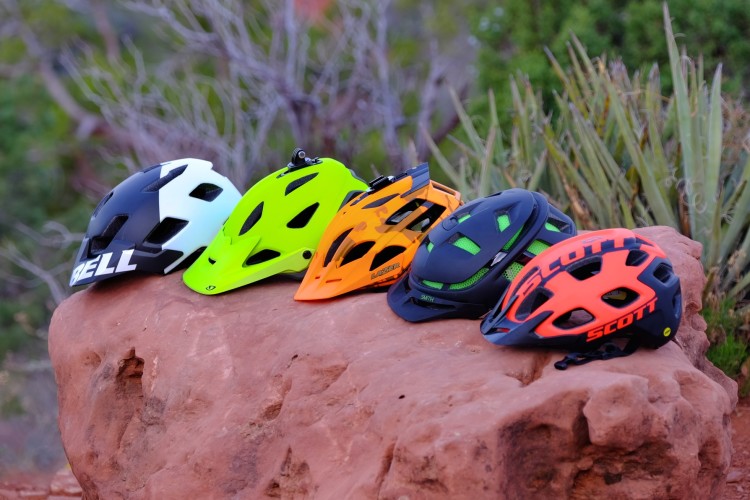 A row of five colorful bicycle helmets displayed on a rocky surface, featuring various styles and brands. The helmets include designs in black and white, bright green, orange, and red with black accents, showcasing a variety of ventilation openings and shapes against a natural background of shrubs and trees.