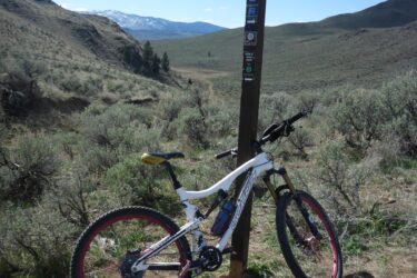 A mountain bike resting against a trail sign in a wide-open landscape. In the background, rolling hills and a distant snow-capped mountain under a blue sky with scattered clouds. The ground is a mix of dirt and sparse vegetation, typical of a rugged outdoor trail environment. Lazy Boy, Sidewinder, Kow-a-Been-Here, Big Horn Loop, Sidewinder, Lazy Boy mountain bike trail.