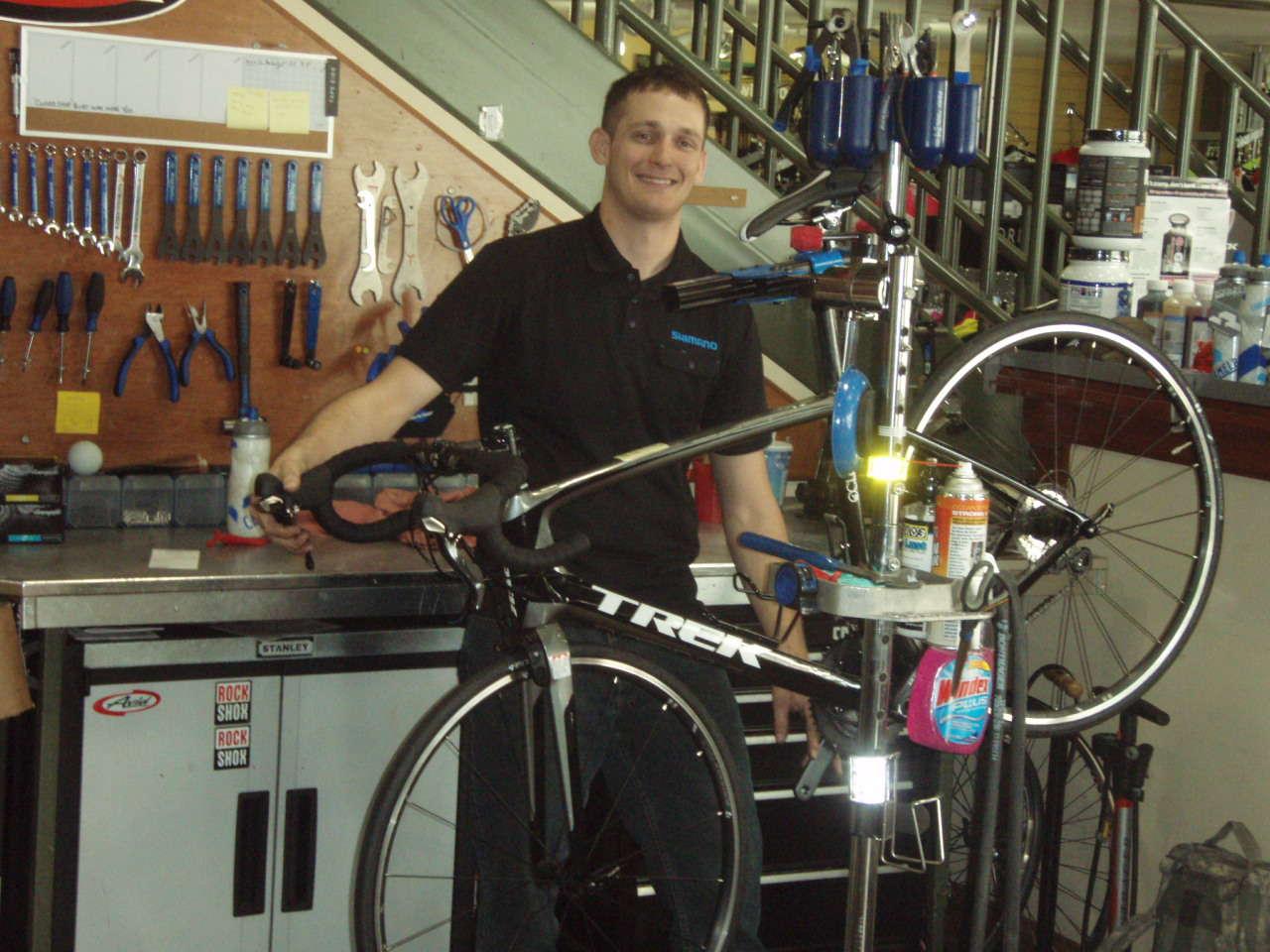 A mechanic smiling while working on a black Trek bicycle in a bike shop, with various tools and equipment visible on the wall and workbench behind him.