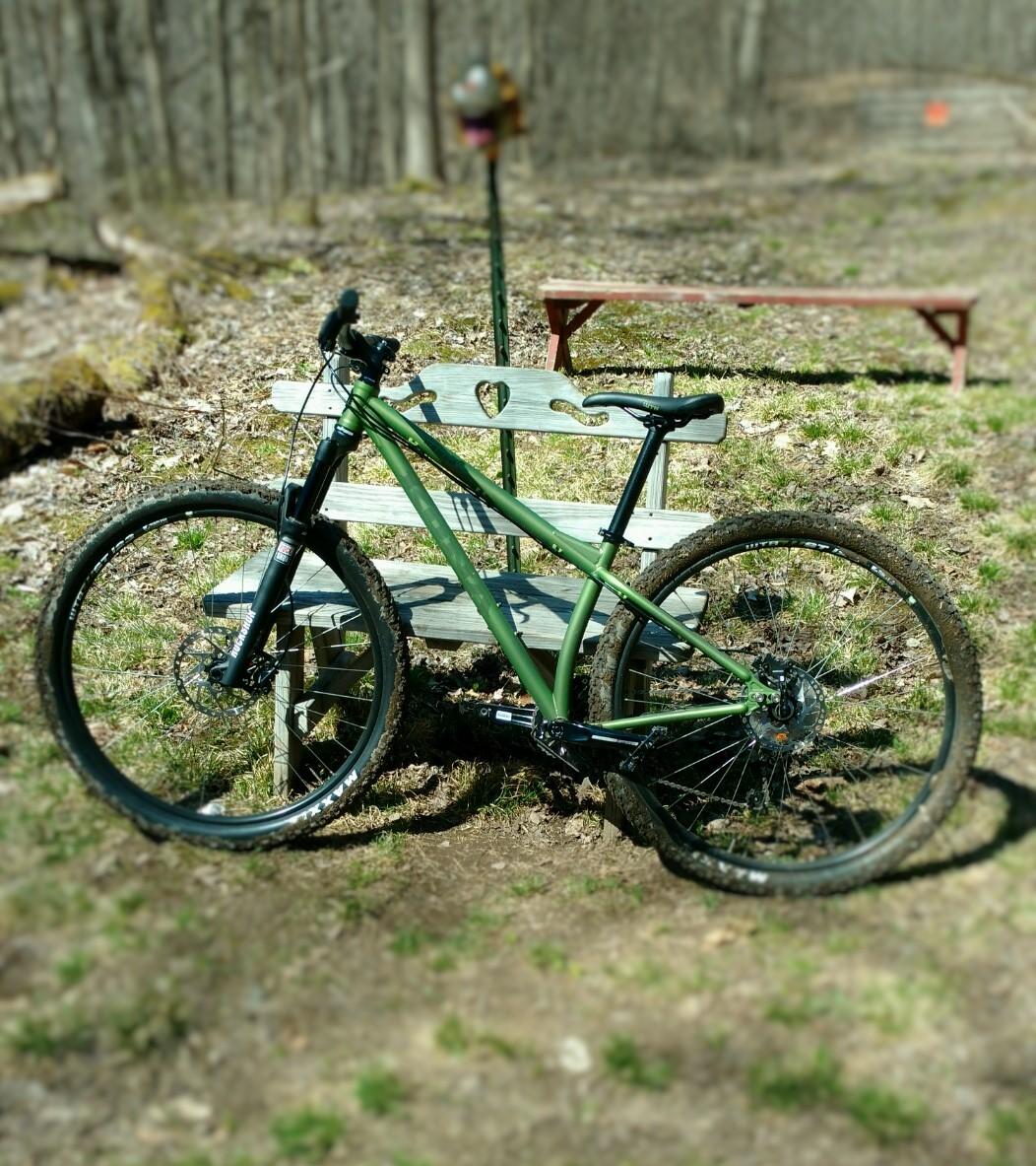 A green mountain bike rests on a wooden bench with a heart-shaped cutout in a wooded area. The ground is covered with dry leaves and grass, and a small table is visible in the background. Sunlight filters through the trees, creating a peaceful, natural setting. Great Bear mountain bike trail.