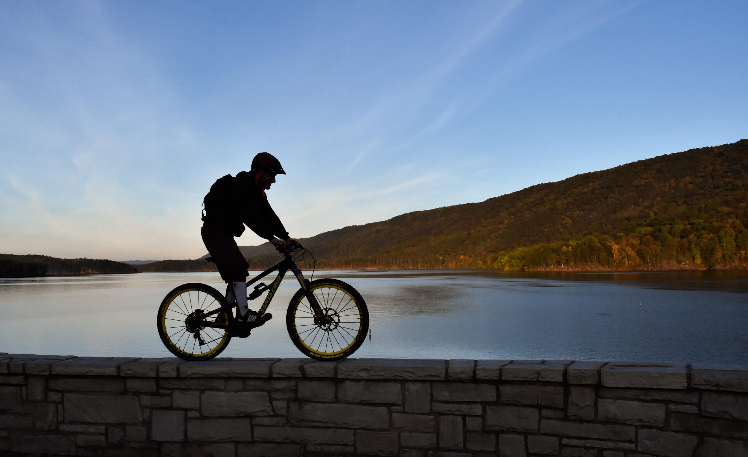 Santa Cruz Nomad Carbon: A silhouette of a person riding a mountain bike along a stone wall, with a calm lake and rolling hills in the background. The sky is clear, displaying soft hues of blue and orange from the setting sun.
