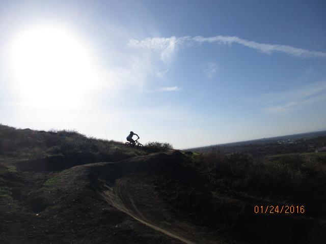 A silhouette of a mountain biker riding on a dirt trail against a bright sunlit sky, with a panoramic view of the landscape below. Irvine Ranch Open Space mountain bike trail.