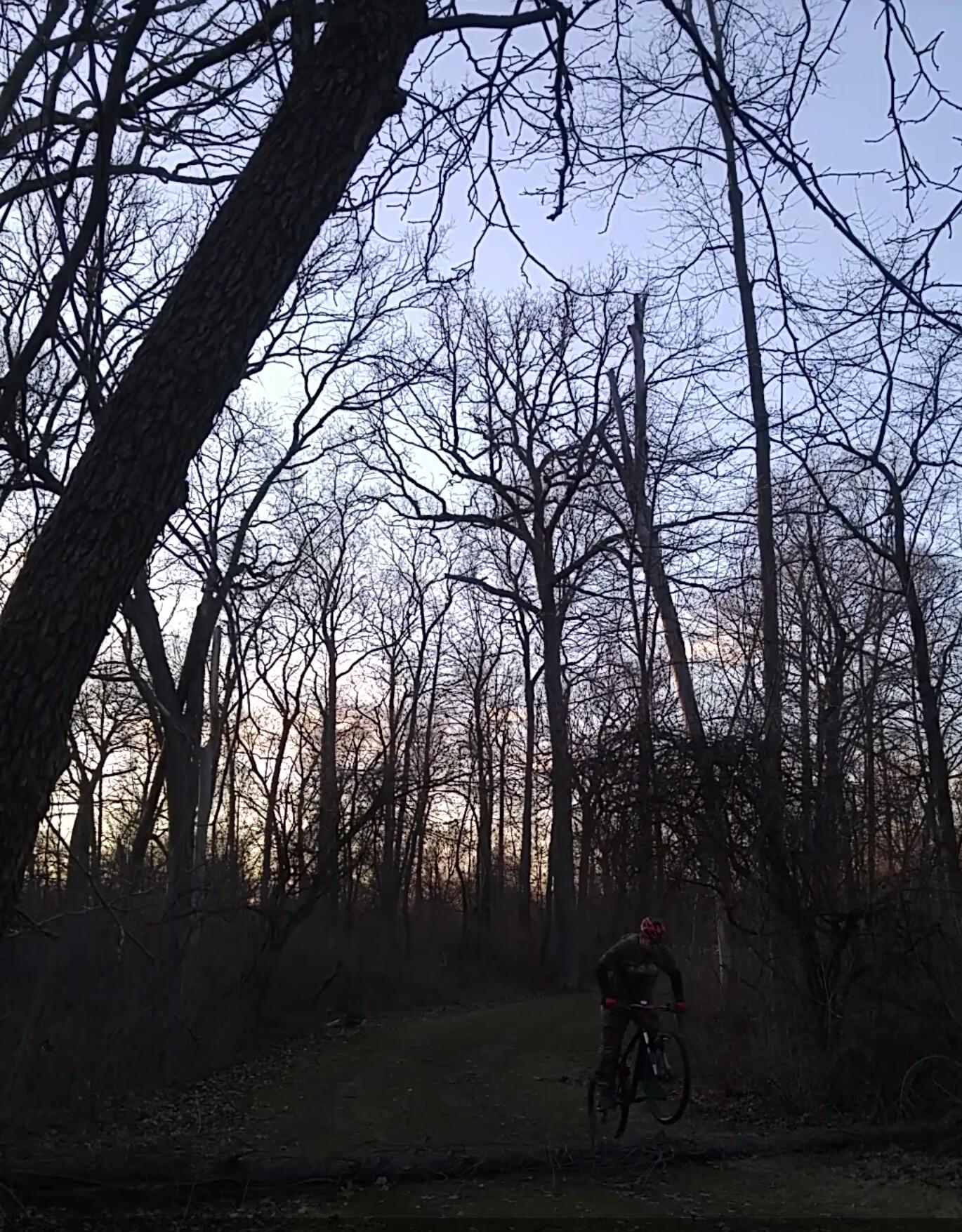 A cyclist in a red helmet rides a mountain bike on a wooded trail surrounded by bare trees at dusk. The sky is a soft gradient of dusk colors as the sun sets in the background. Elizabeth Park Clydesdale Loop mountain bike trail.