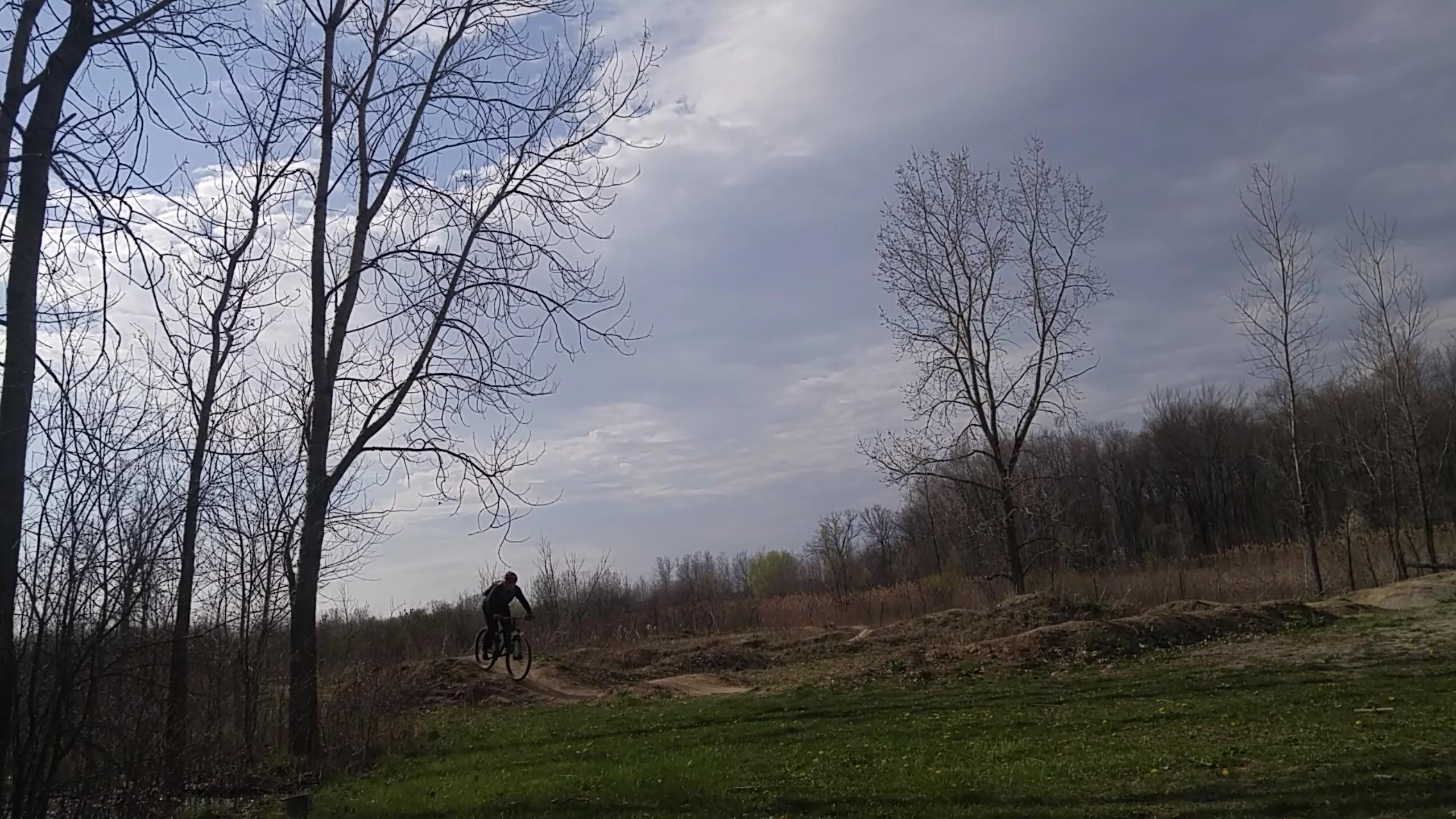 A mountain biker performing a jump on a dirt trail in a rural area, surrounded by bare trees and a cloudy sky. Morton-Taylor Trail mountain bike trail.
