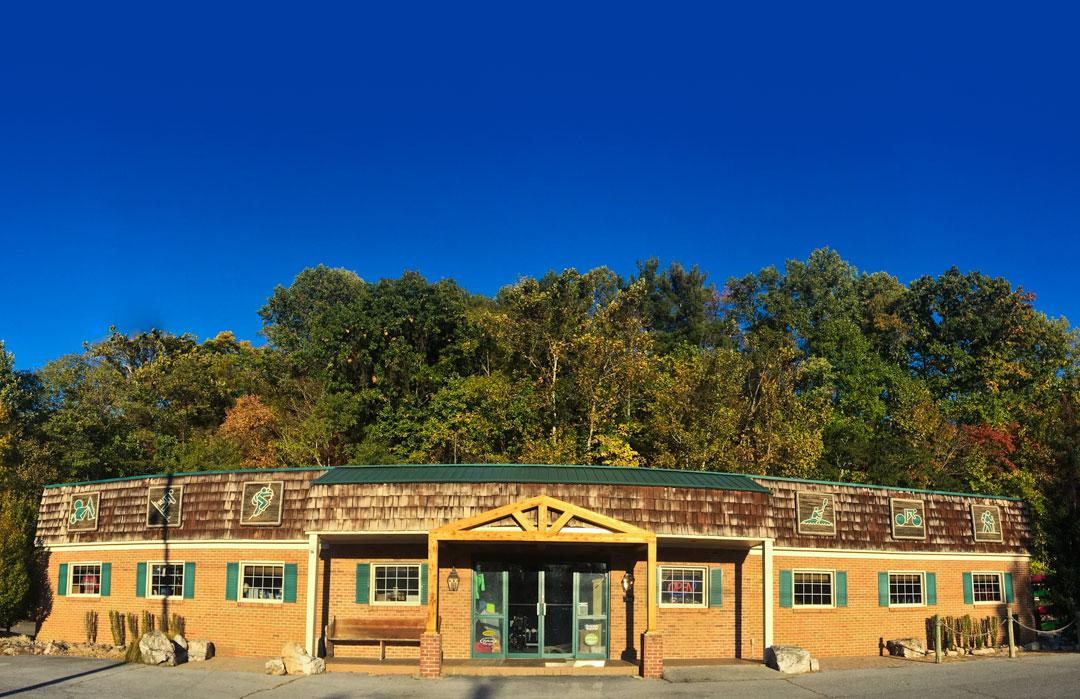 A brick building with a wooden awning and green trim, set against a backdrop of colorful trees. The building features large windows and decorative signs on the roof depicting various outdoor activities. The sky is clear and blue.