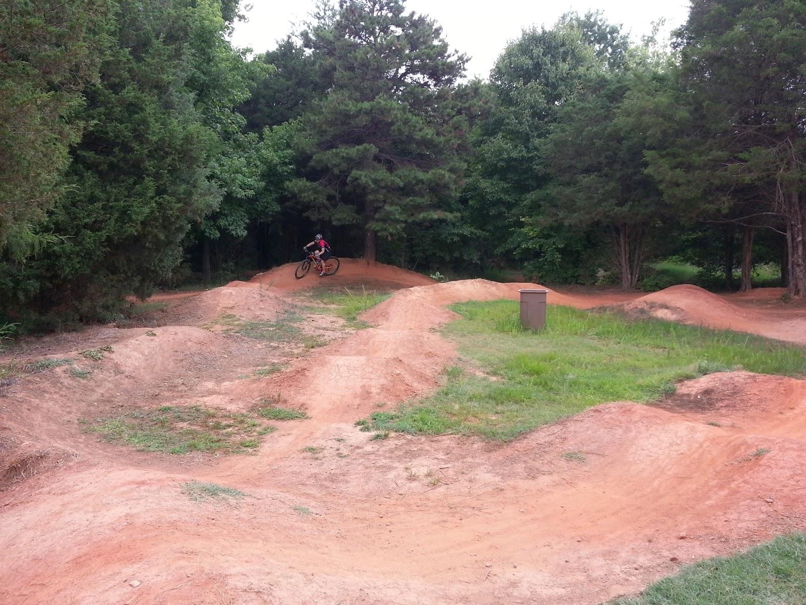 A cyclist riding a mountain bike on a dirt track with jumps and bumps, surrounded by trees and greenery. The track is made of reddish dirt, with patches of grass visible. Signal Hill mountain bike trail.