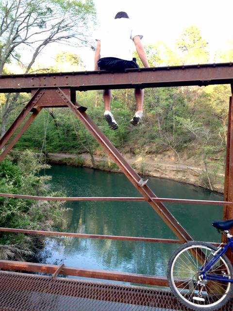 A person sitting on the edge of a metal bridge, overlooking a calm body of water below, surrounded by greenery. A blue bicycle is resting against the bridge railing. Spadra Creek Nature Trail mountain bike trail.