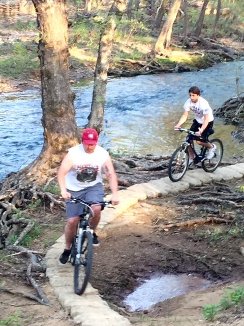 Two young men riding bicycles on a dirt trail alongside a stream in a wooded area. One rider is wearing a red cap and a white sleeveless shirt, while the other is in a white t-shirt and black shorts. The setting features trees and a natural landscape, with the stream visible in the background. Spadra Creek Nature Trail mountain bike trail.