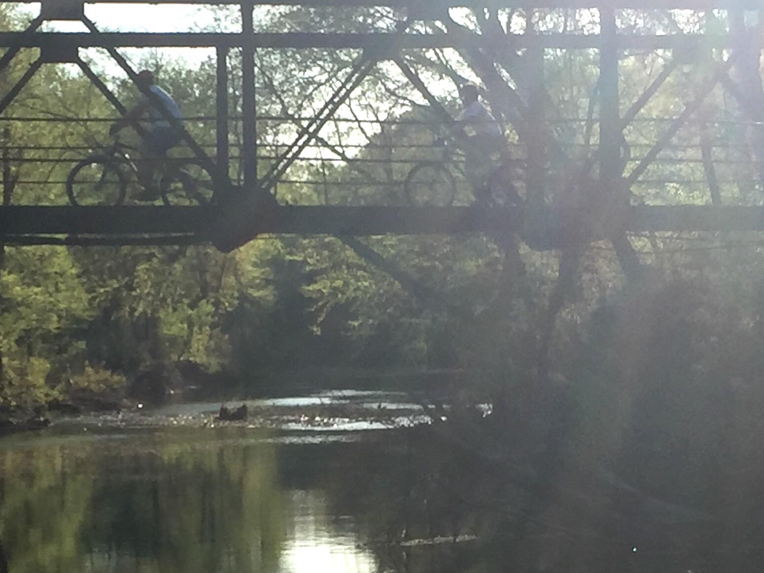 Two cyclists riding along a metal bridge over a calm river, surrounded by lush greenery and soft sunlight filtering through the trees. Spadra Creek Nature Trail mountain bike trail.