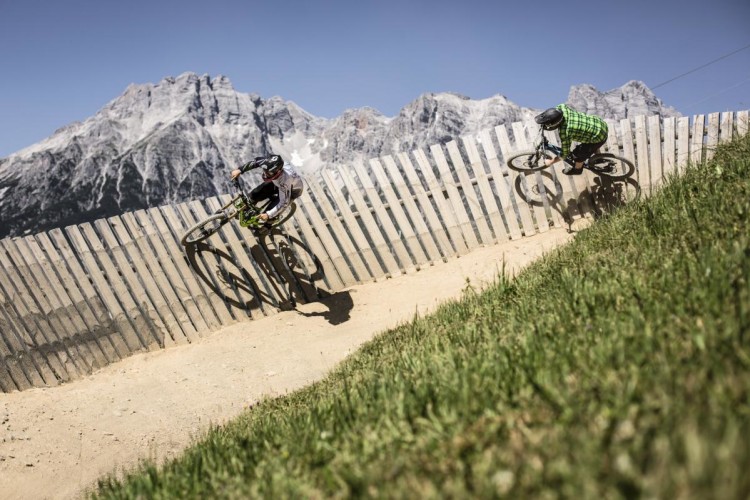 Two mountain bikers maneuvering along a curved wooden track with a backdrop of large, rocky mountains under a clear blue sky. One rider is leaning into the turn while the other is slightly behind, both showcasing dynamic action in a natural outdoor setting.