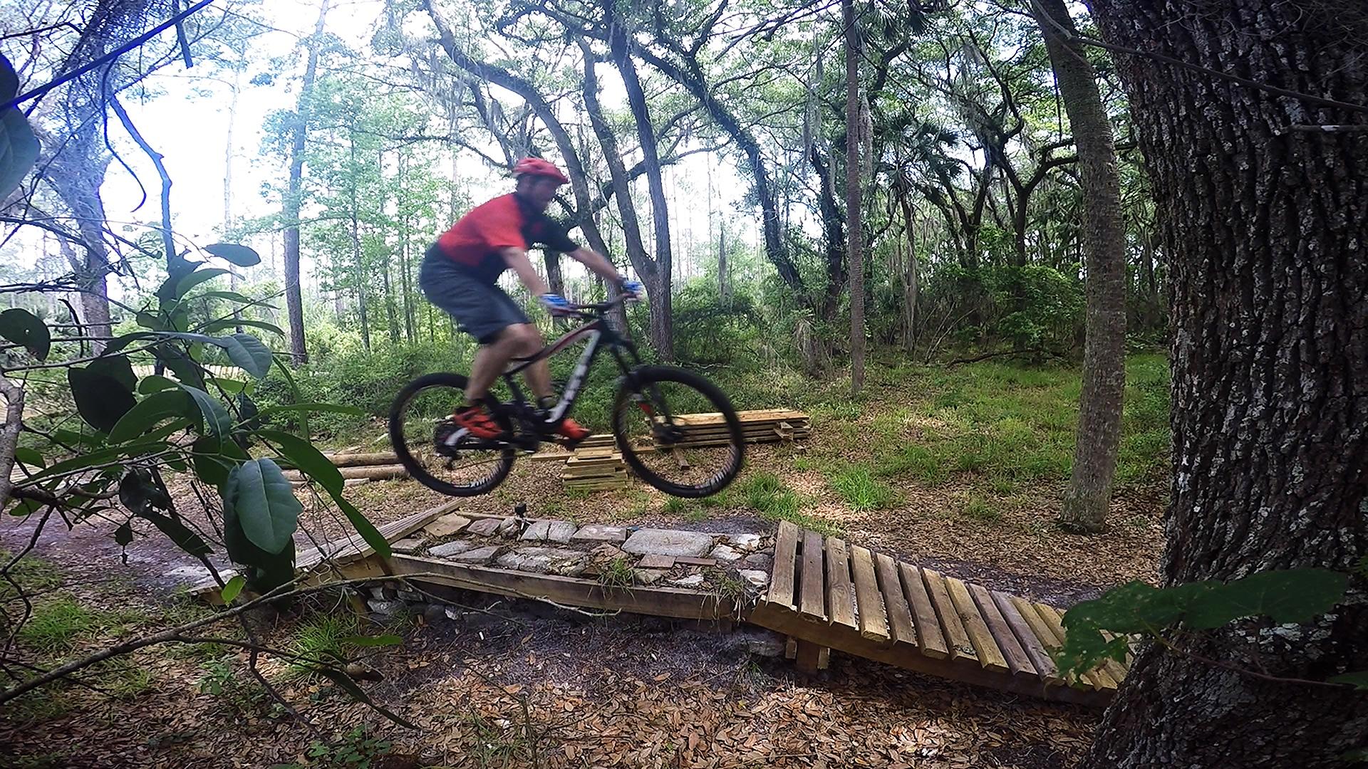 A mountain biker in a red shirt and helmet jumps over a wooden ramp while riding through a forested trail, surrounded by green trees and foliage. Nocatee mountain bike trail.