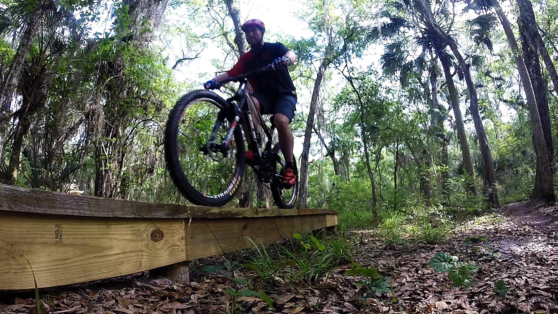 A mountain biker performing a jump off a wooden bridge in a lush forest setting. Trees and greenery surround the trail, with scattered leaves on the ground. The biker is wearing a helmet, gloves, and casual cycling attire. Nocatee mountain bike trail.