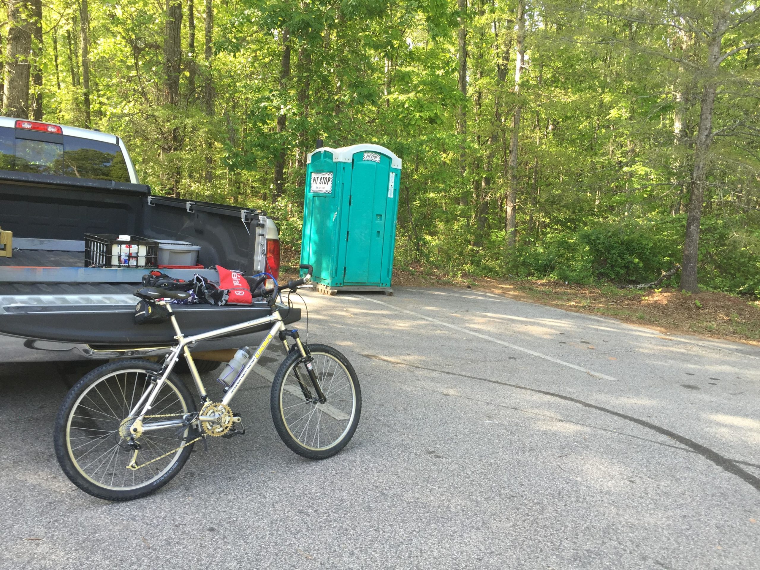 A silver mountain bike is parked next to the open tailgate of a pickup truck, which contains various supplies. In the background, a portable restroom is visible near a tree line, and the scene is set in a wooded area with green foliage. The Trails At The Beach mountain bike trail.