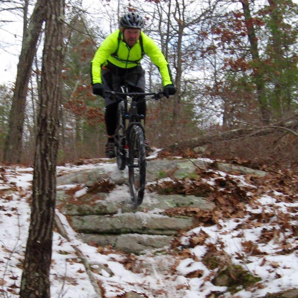 A mountain biker wearing a bright yellow jacket is navigating over rocky terrain covered with snow and fallen leaves in a wooded area. Trees surround the path, and the biker is captured mid-jump, demonstrating skill and balance on the bicycle. Vietnam Trails mountain bike trail.
