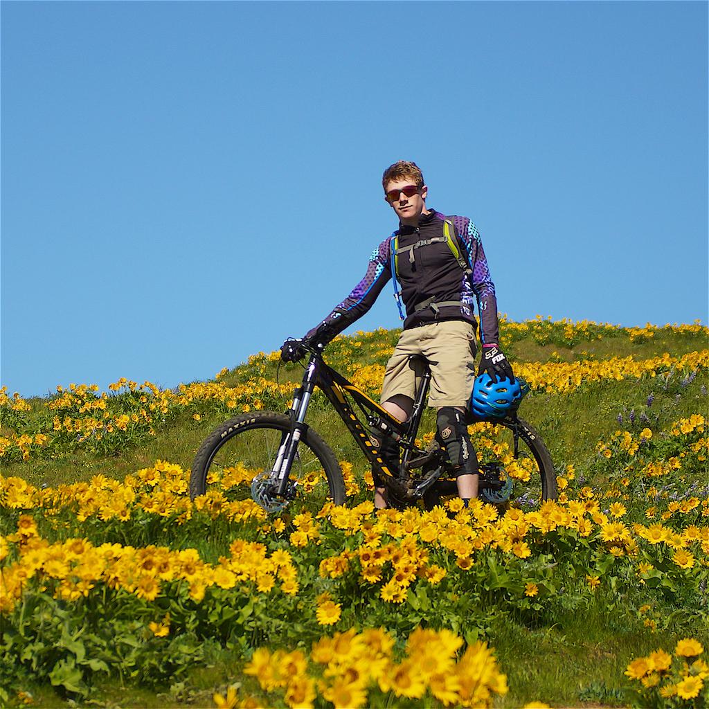 A young mountain biker stands beside his bike on a hillside covered with vibrant yellow flowers under a clear blue sky. He is wearing a helmet, sunglasses, and protective gear, and is posing confidently in the colorful landscape. Syncline mountain bike trail.
